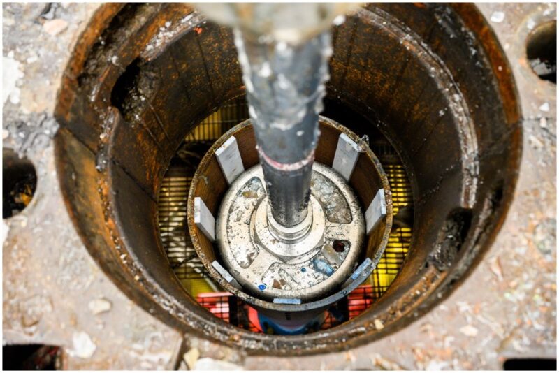 Looking down into a borehole lined with rusty metal, a shiny metal rod attached to a shiny cylinder in the center of the borehole.